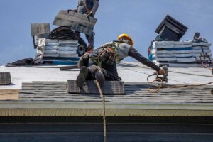 Workers remove old shingles from a residential roof while exposed roof decking is inspected.
