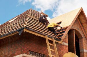 Person carefully using a secured ladder beside a dry asphalt-shingle roof with safety equipment nearby.